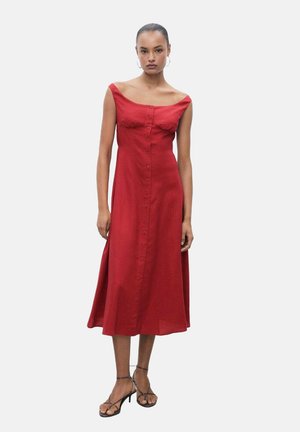 Robe en lin rouge avec un design sans épaule, des boutons sur le devant, une jupe fluide et une fente sur le côté. Associée à des sandales à lanières.