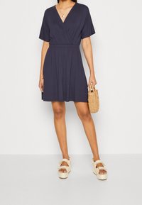 Granatowa krótka sukienka z dekoltem w kształcie litery V, krótkimi rękawami i elastyczną talią. Połączona z kremowymi sandałami na koturnie i plecioną torebką.