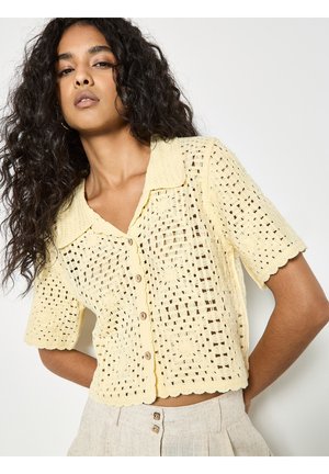 Donna con capelli ricci che indossa una maglia all'uncinetto giallo pallido a maniche corte con bottoni e pantaloni beige a vita alta su sfondo bianco.