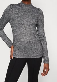 Haut gris côtelé à manches longues avec col montant, présentant des manches légèrement bouffantes et un motif texturé, associé à des leggings noirs.