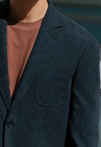 Marineblå blazer med tekstur og et subtilt blomstermønster, enkelt knaplukning og en brystlomme, lagt over en rustfarvet skjorte.