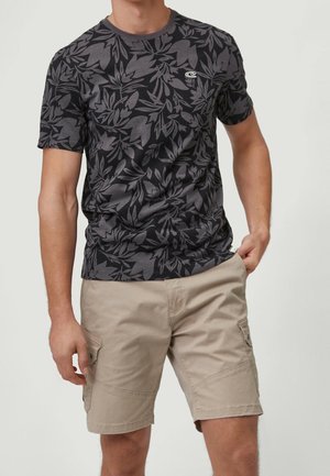 Uomo che indossa una maglietta nera e grigia con motivo a foglie e pantaloncini cargo beige con tasche, in piedi contro uno sfondo semplice.