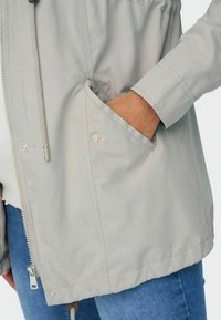 Světle šedá větrovka se zipem vpředu, kapucí na stahování a bočními kapsami. Má hladkou texturu a mírně oversized střih.