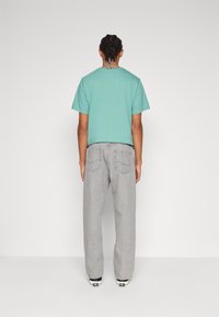 Osoba s pletenými vlasy, oblečená v tyrkysovém tričku s krátkým rukávem, volných šedých džínech a bílých teniskách, stojící čelem k obyčejnému bílému pozadí.