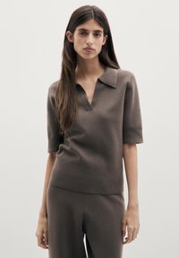 Giovane donna con capelli lunghi e scuri che indossa una maglia lavorata a maglia taupe a maniche corte con colletto e pantaloni abbinati, in piedi davanti a uno sfondo semplice.