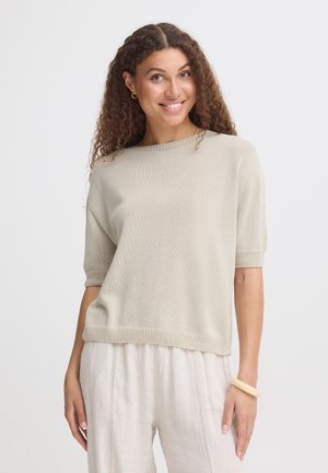 Kvinde med krøllet hår smiler og har en beige kortærmet strik top på, lyse bukser, små guldringede øreringe og et beige armbånd.