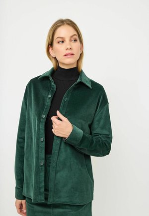 Green corduroy shirt jacket with a button front, spread collar, and long sleeves, displayed over a black ribbed top. Smooth texture.