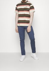 Koszulka w paski w kolorach kremowym, czerwonym, zielonym i pomarańczowym, zestawiona z niebieskimi spodniami o dopasowanym kroju. Model nosi białe trampki, stoi na neutralnym tle.