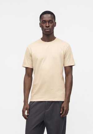 Junger Mann, der frontal steht und ein schlichtes beiges Kurzarm-T-Shirt sowie dunkelgraue Hosen vor einem weißen Hintergrund trägt.