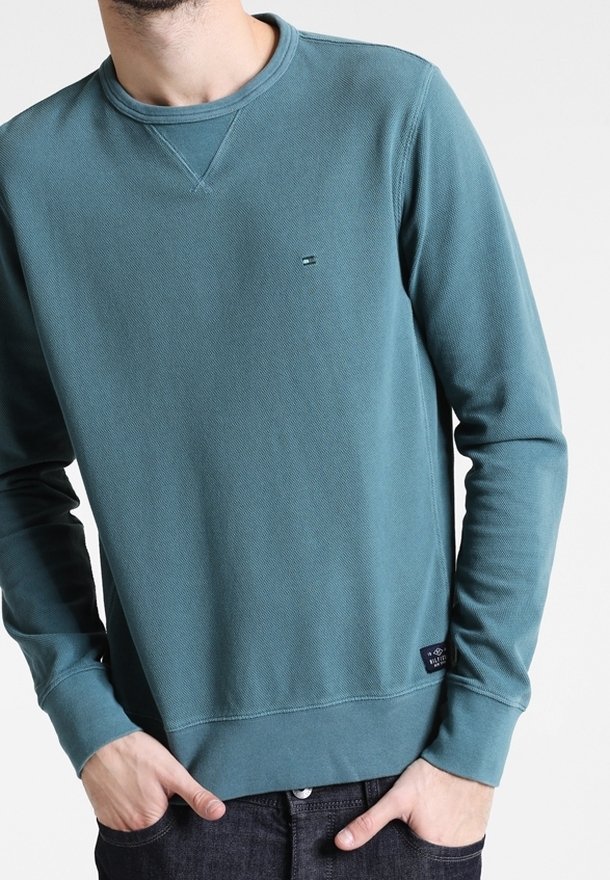 Homme portant un sweat-shirt col rond bleu sarcelle à manches longues avec un petit logo sur la poitrine et les mains dans les poches, sur fond blanc.