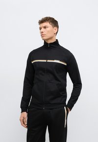 Μαύρο μπουφάν με φερμουάρ, κατασκευασμένο από απαλό ύφασμα, με χρυσή και λευκή γραμμή στο στήθος και λογότυπο "BOSS". Δύο μπροστινές τσέπες.