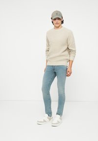 Maglione in misto beige, jeans attillati blu chiaro e sneakers bianche con dettagli grigi. Il modello indossa un cappello beige, mostrando un look casual.