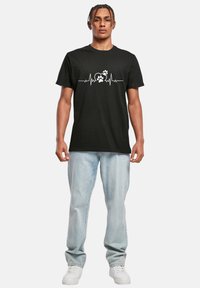 Schwarzes Baumwoll-T-Shirt mit einem weißen Herzschlaglinien-Motiv und einem Pfotenabdruck-Graphic. Kombiniert mit hellblauen Jeans und weißen Turnschuhen.