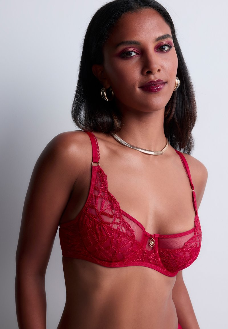 Soutien-gorge en dentelle rouge avec des panneaux en maille, des motifs géométriques, des bretelles réglables et un petit accent doré au centre. Matériau léger.