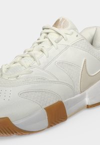 Άσπρο παπούτσι Nike με μπεζ λογότυπο, υφασμάτινη υφή, δερμάτινες επικαλύψεις, λευκά κορδόνια και λεπτομέρεια καφέ λαστιχένιας σόλας.