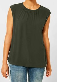 Tmavě zelený bezrukávový top s nabíraným výstřihem a ozdobnými cvočky na ramenech, spárovaný se světle modrými džínami. Hladká textura látky.