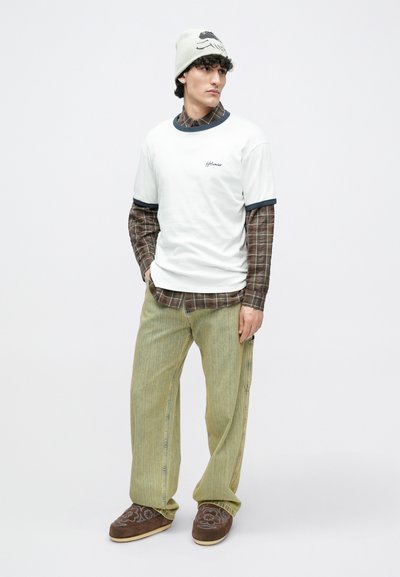 Fehér póló tengerészkék szegéllyel, barna kockás gombos ing felett, laza zöld kordbársony nadrággal és hímzett barna papuccsal párosítva.