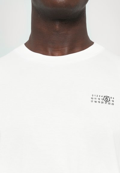 T-shirt de algodão branca com decote redondo, apresentando um pequeno design impresso em preto na área do peito esquerdo, exibindo números e o número 6.