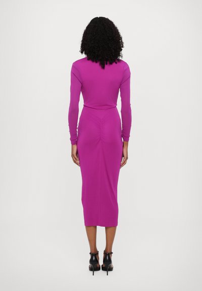 Φόρεμα μακρυμάνικο, εφαρμοστό σε ζωηρό ροζ χρώμα με πτυχώσεις στην πλάτη, συνδυασμένο με μαύρες ψηλοτάκουνες γόβες με λουράκι στον αστράγαλο. Λείο υλικό.