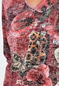 Czerwony wzorzysty top z motywami kwiatów i motyli, ozdobiony kryształkami, z dekoltem w serek. Materiał wydaje się miękki o fakturowanym wykończeniu.