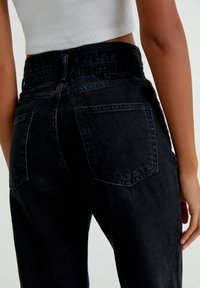 Femme portant un jean en denim noir taille haute et un haut court blanc côtelé, visible de la taille aux jambes, avec une main détendue le long du corps.