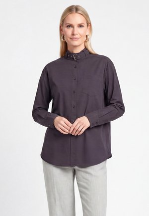 Frau mit langen blonden Haaren, die ein dunkles Hemd mit Knöpfen und einem Schnallkragen sowie helle Hosen trägt, stehend mit verschränkten Händen.