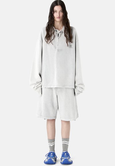 Personne aux cheveux longs portant un sweat-shirt gris clair surdimensionné et un short, des chaussettes grises et des baskets bleues, debout devant un fond blanc.