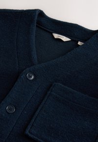 Tume sinine nööbitav kardigan rinnatasku ja kaeluse siseserval oleva valge suuruse sildiga "M".