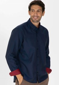 Chemise boutonnée bleu marine à manches longues avec des accents rouges sur les poignets. Elle possède un col classique et une texture lisse.
