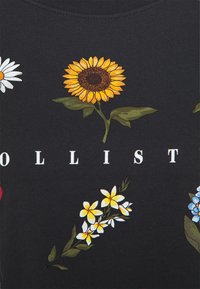 Černá látka s barevnými květinovými potisky, včetně slunečnic, kopretin a modrých květů, s nápisem "OLLIST" bílými písmeny.