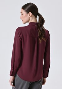 Burgundy long-sleeve blouse with a high collar, pleated back detail, and button cuffs, made from smooth fabric.