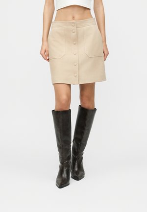 Kvinne iført en beige skjørt med knapper foran og frontlommer, samt svarte skinnstøvler til knærne, stående mot en ensfarget bakgrunn.