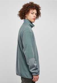 Fleece pullover vaaleansinisen sävyssä, jossa on korkea kaulus ja vetoketju. Harmaat hihat tarjoavat tyylikästä tekstuurikontrastia. Tavallinen istuvuus ja kaareva helma.