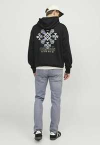 Sweatshirt à capuche noir avec un motif géométrique bleu et blanc au dos, porté avec un jean gris clair et des baskets noires.