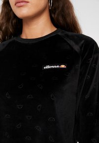 Acercamiento de una persona que lleva un sudadera negra de Ellessee con un sutil patrón en relieve y un pequeño logo en el pecho.