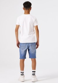 Camiseta de algodón blanca, pantalones cortos de mezclilla azules, zapatillas blancas y calcetines a rayas en blanco y negro. Vista trasera, con costuras visibles y detalles de bolsillo.
