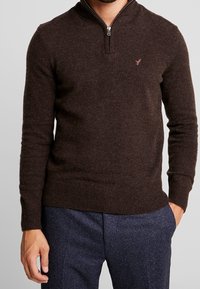 Brun zip-neck tröja med ribbade ärmslut och nederkant, tillverkad av strukturerad stickad tyg, med en liten logobrodering på bröstet.