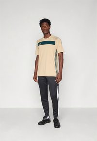 Beige trenings-T-skjorte med grønn horisontal stripe og tre grønne vertikale striper på siden; kombinert med svarte treningsbukser med hvite sidestreker.