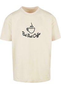 Cremefarvet bomulds T-shirt med sort tekst "Men først kaffe" og et kaffekops grafik trykt på fronten. Korte ærmer, rund hals.