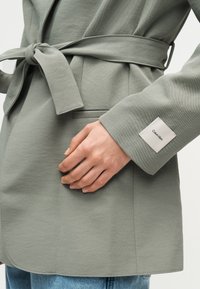 Blazer gris ceinturé avec un patch Calvin Klein sur le poignet, porté sur un jean bleu, main reposant près d'une poche avant.
