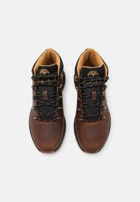 Botas de senderismo de cuero marrón con parte superior de malla negra, acentos naranjas y ojales de metal. Presenta un collar acolchado y un logotipo en la lengua.