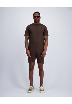 Man wearing brown t-shirt and matching shorts, beige sneakers, sunglasses, and a wrist bracelet, standing against a plain light background.