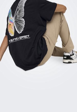 Person sitting wearing black T-shirt with colorful butterfly graphic and "The Butterfly Effect" text, beige pants, and black-and-white sneakers.