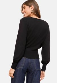 Femeie zâmbind, purtând cercei triunghiulari din aur, un pulover negru cu mâneci lungi și manșete și blugi de culoare albastră închisă, văzută din spate.
