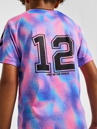 Barn iført en pink og blå tekstureret kortærmet skjorte med sort nummer 12 og teksten "Light Up The World" på ryggen.