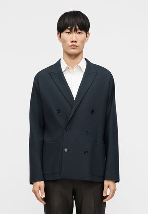 Giacca doppiopetto blu navy con rever a lancia, quattro tasche frontali e tessuto dalla trama liscia, abbinata a una camicia bianca e pantaloni marroni con trama distinta.