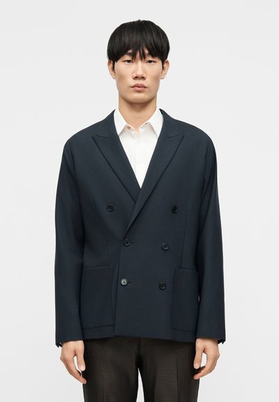 Marineblaue zweireihige Jacke mit Reverskragen, vier vorderen Taschen und glatter Stoffstruktur, kombiniert mit einem weißen Hemd und braunen strukturierten Hosen.