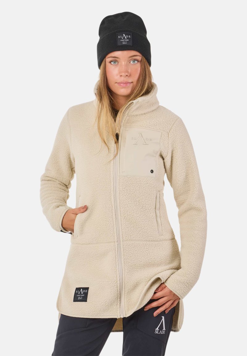 Giacca in pile beige con colletto alto, tasche laterali e tasca sul petto. Presenta il logo del brand e una superficie texturizzata. Accentuata da un berretto nero.