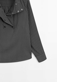 Chemise à manches longues gris foncé avec fines rayures verticales noires, boutonnée à l'avant partiellement déboutonnée, avec boutons-pression sur le col et la poitrine.