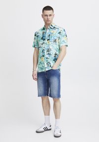 Camicia a maniche corte di colore azzurro chiaro con stampa floreale tropicale, abbinata a pantaloni corti di jeans. Footwear con tre strisce nere su base bianca.